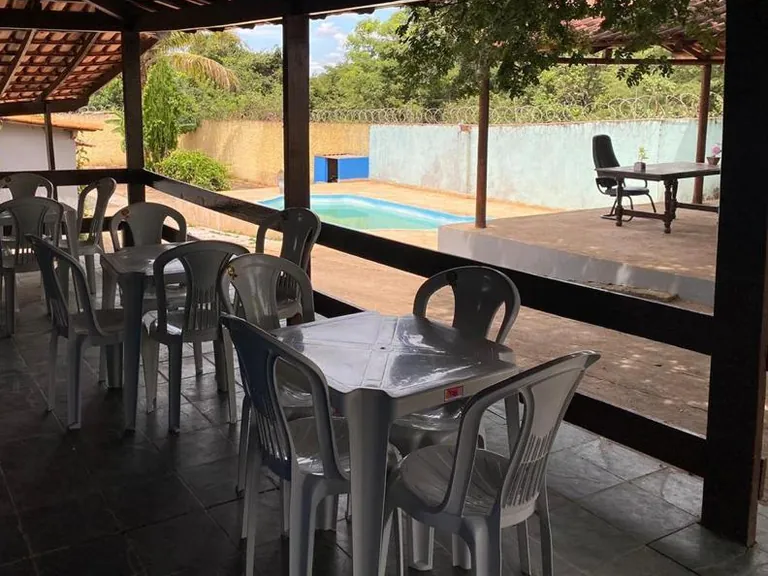 Área coberta com telhado de madeira, mesas e cadeiras de plástico brancas e piscina ao fundo