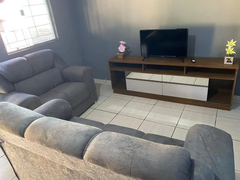 Sala com dois sofás cinza, rack de madeira com televisão desligada e dois vasinhos de flores